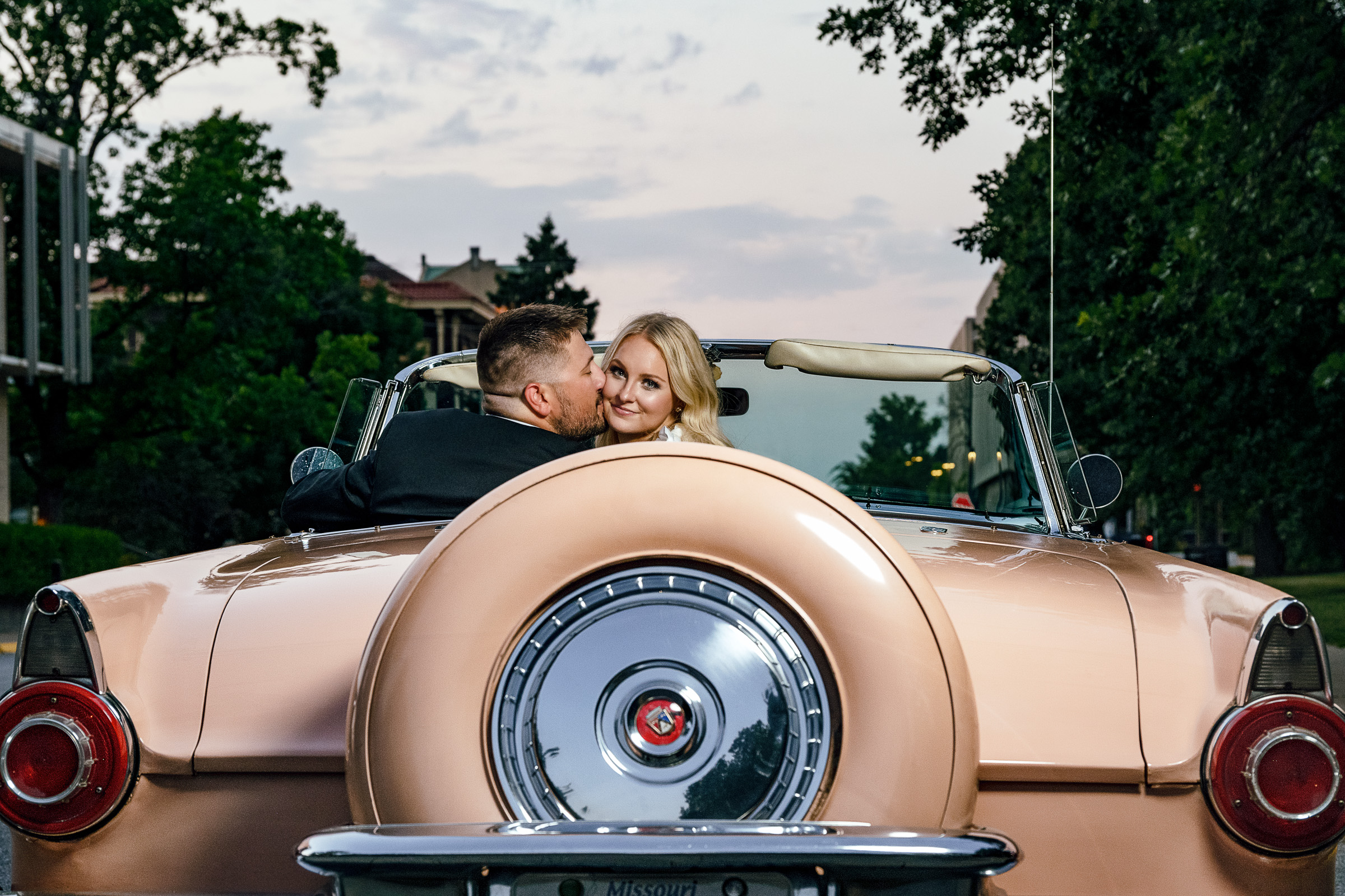 engaged couple sitting in 1956 buckskin colored thunderbird convertible while guy kisses woman on cheek