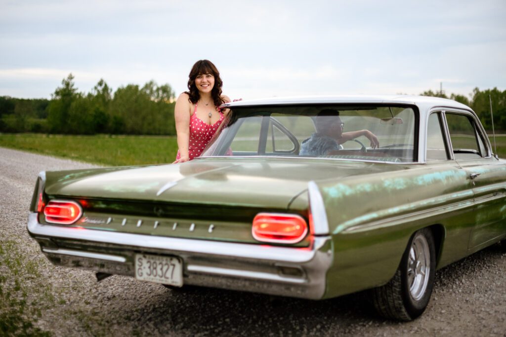 Brunette woman wearing pink dress with white polka dots standing next to green car.