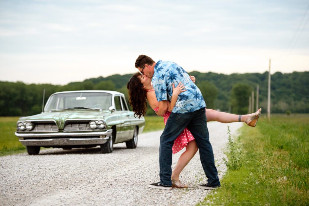 Engaged couple kiss on country road with green car in the background.