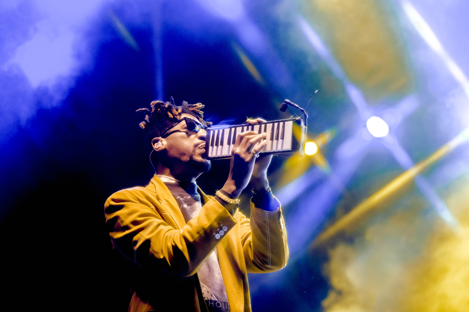 John Batiste plays melodica while blue and yellow lights create a star burst in background.