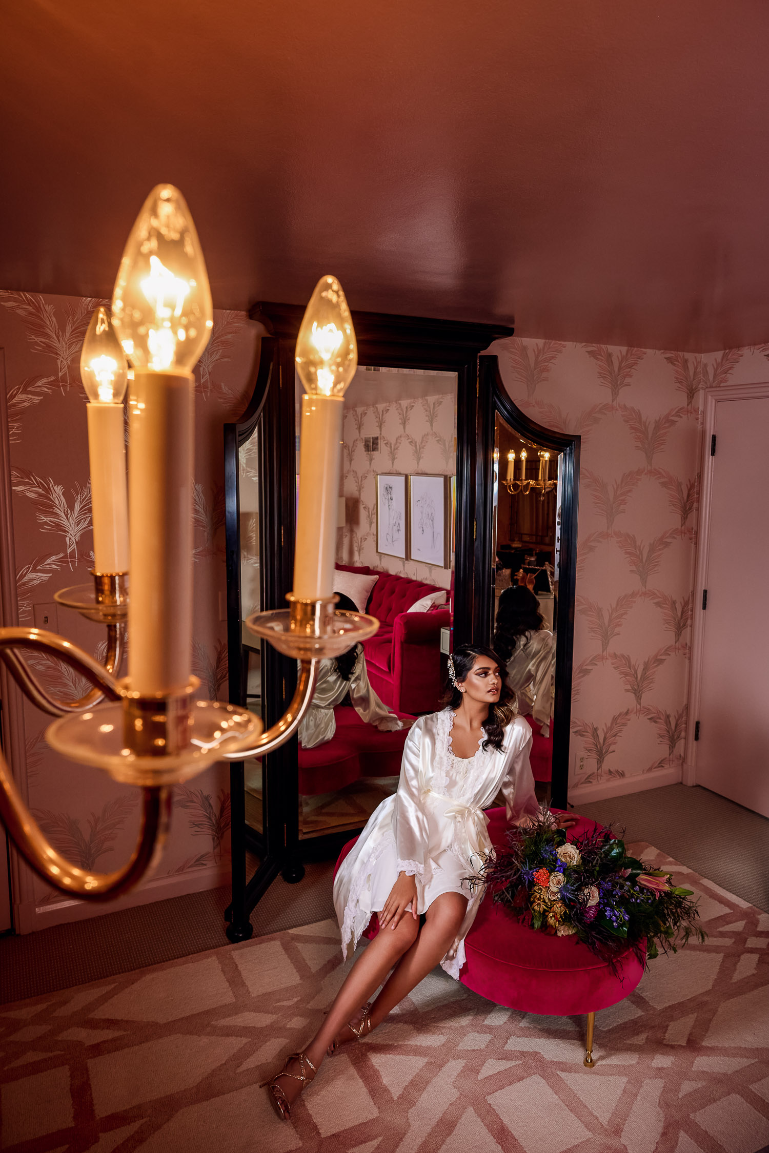Bride wearing silk robe lounges in front of mirror and chandelier in pink room.