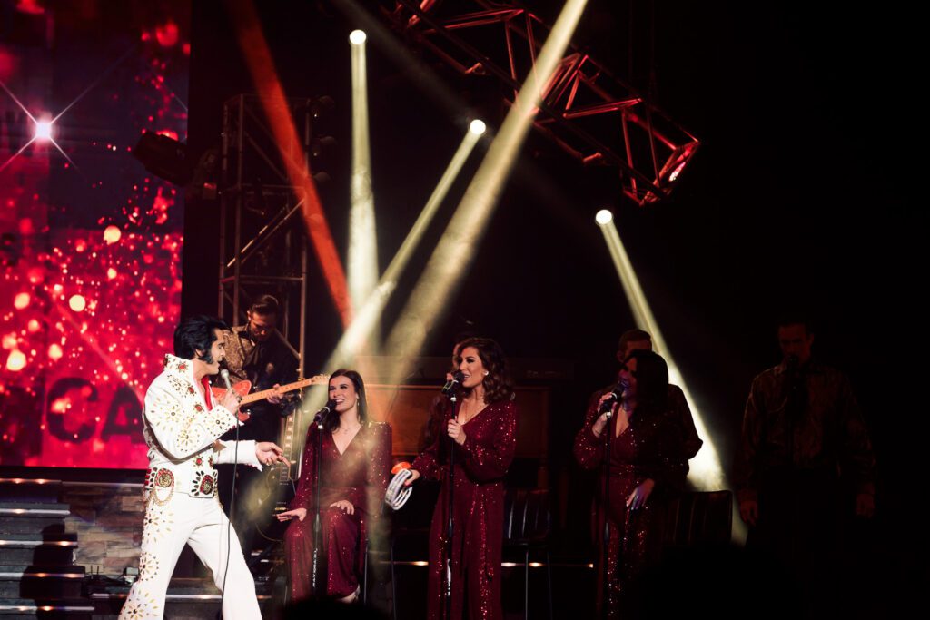 Elvis tribute artist singing on stage with three lady back up singers wearing red sequin dresses.