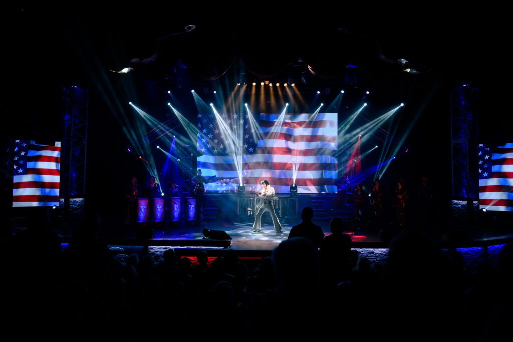 Dean Z performs as Elvis Presley with American Flags on screens.