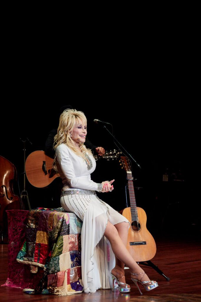 Dolly Parton sits on a coat of many colors quilt on stage at the Congress of Country Music