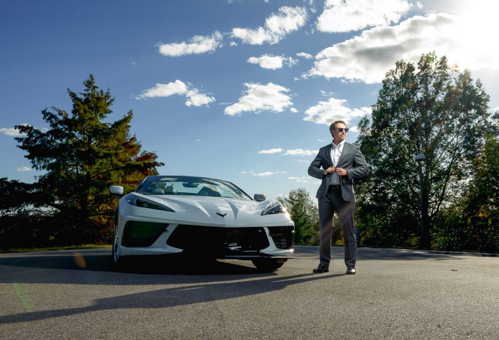 Senior guy wearing suit and sunglasses poses with white corvette