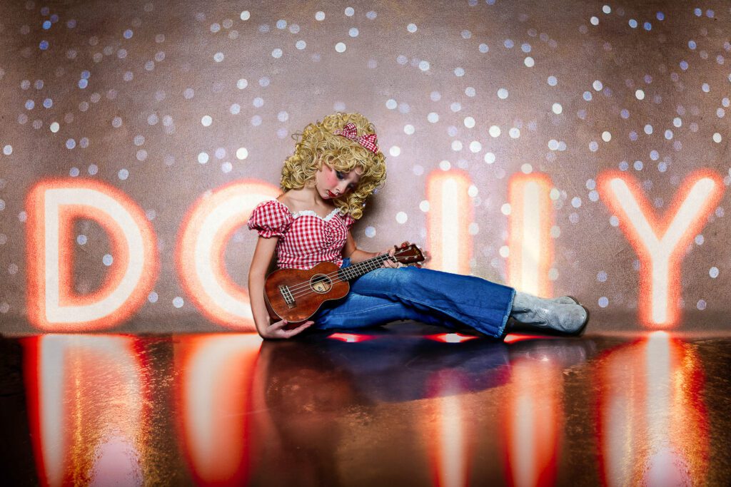 young girl dressed as Dolly Parton sits on the ground with small guitar