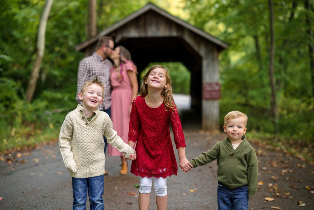 kids hold hands while mom and dad kiss in front of covered bridge