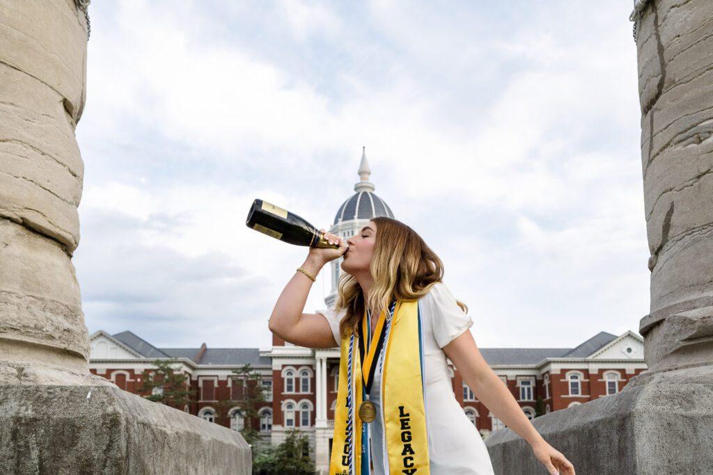 senior graduate woman drinking champagne by mizzou columns